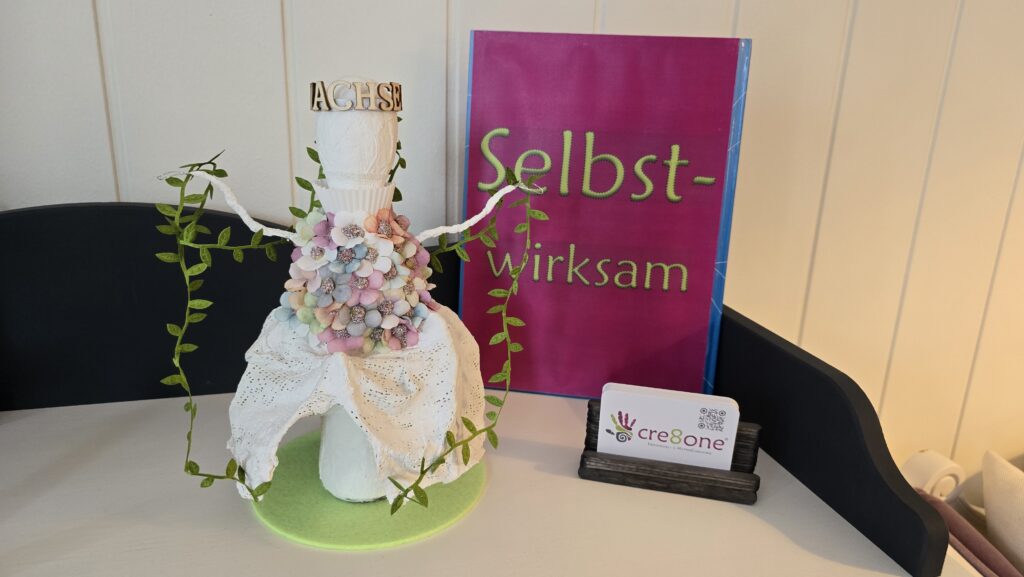 Kleines Kunstobjekt. Eine Figur die den Wachstum darstellt, mit Ranken in der Händen und bunten Blüten am Oberteil eines barocken Kleides. Im Hintergrund ein Buch mit großer Aufschrift "Selbstwirksam".